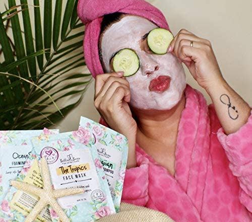 Woman in pink robe with towel turban wearing white mask and cucumber slices beside Bella and Bear Hello Glow face mask packets